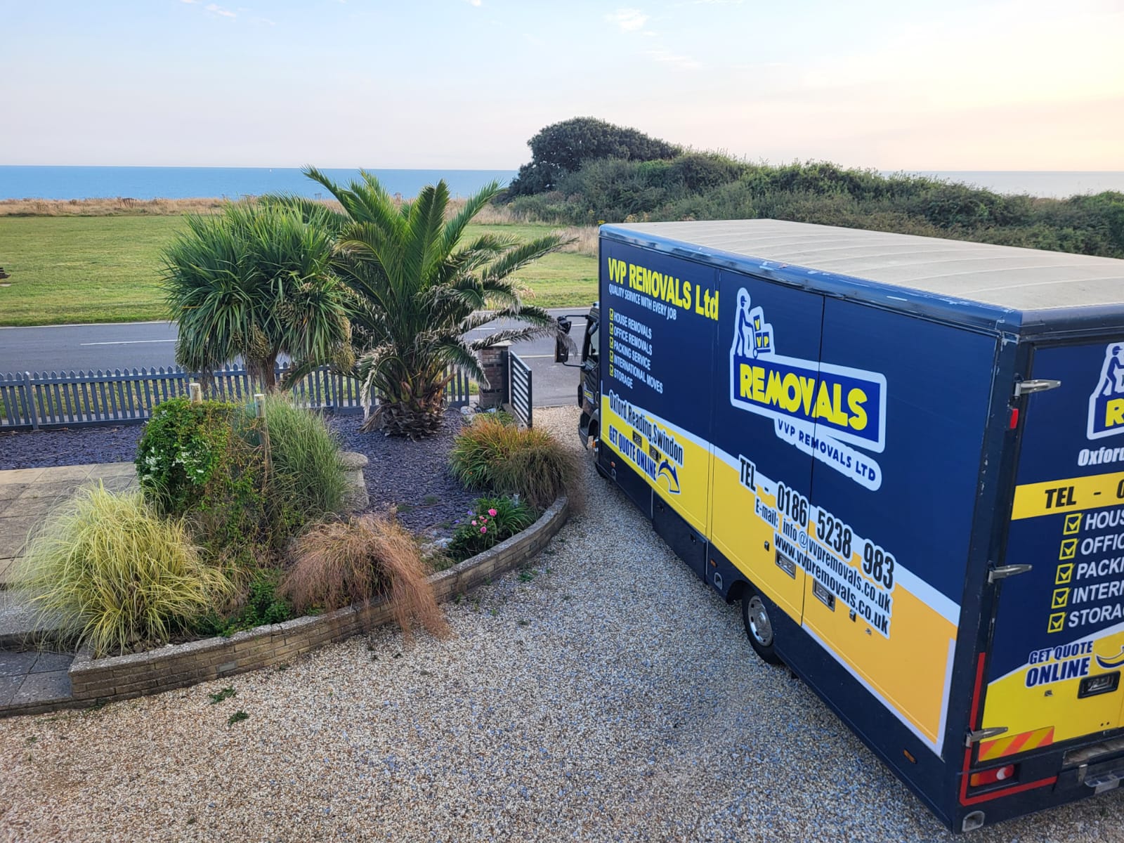A navy and amber VVP lorry beside a coastal garden with palm trees and the sea behind